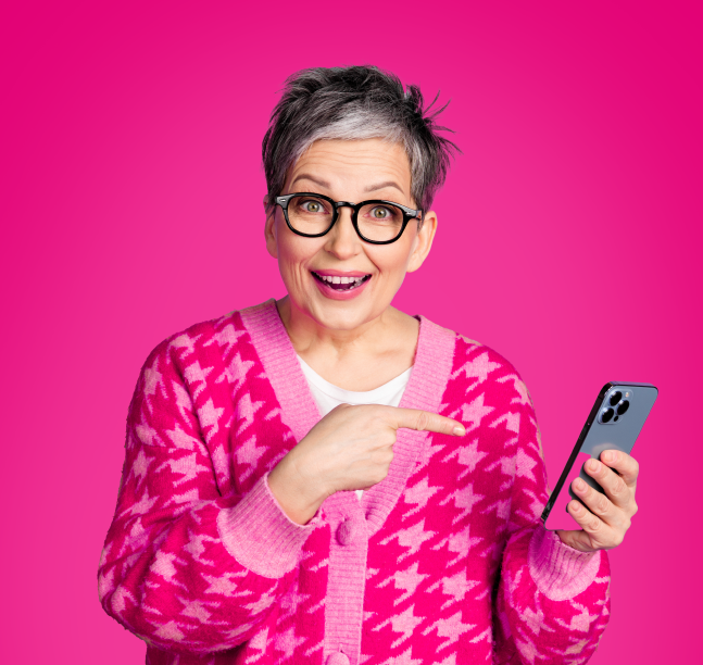 Starsza kobieta w okularach i różowym swetrze uśmiecha się i wskazuje palcem na trzymany w ręku smartfon. Tło jest jednolicie różowe.